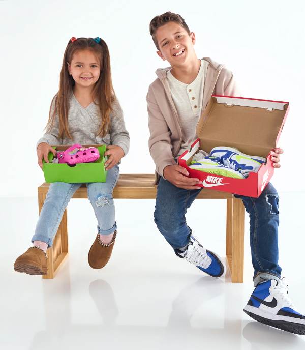 A cute sibling duo are holding Crocs clogs and Nike sneakers in hand are sitting on wooden chair wearing stylish footwear