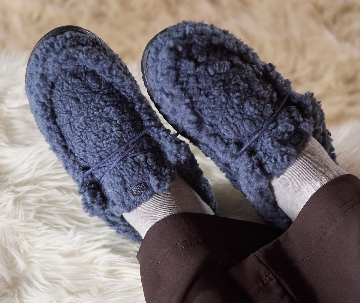 A zoomed in lifestyle image someone wearing blue furry men's HEYDUDE wally slippers inside over a shag rug.