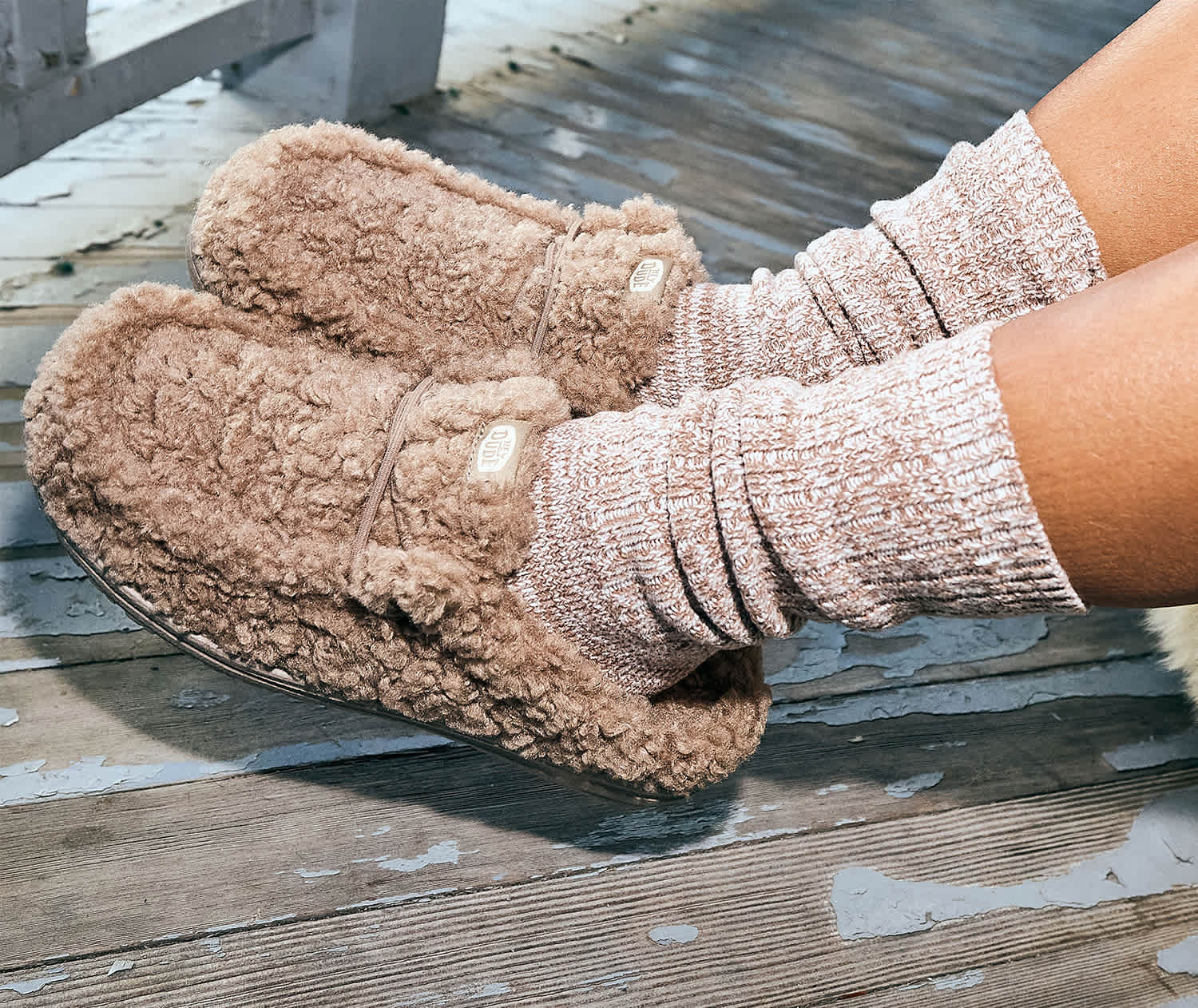 A closeup lifestyle image of brown furry women's HEYDUDE wendy slippers being worn outside on a deck.