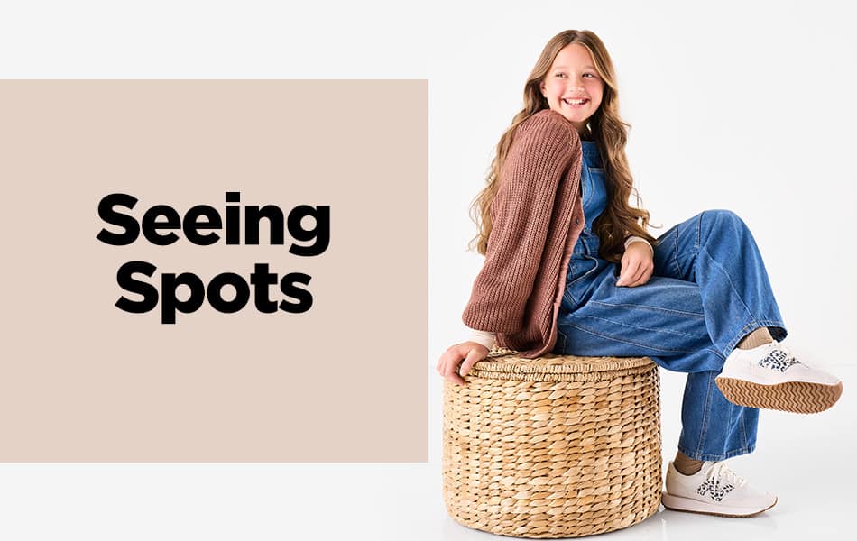 A teenage girl smiling, sitting on a woven ottoman wearing Women's New Balance 370 Sneakers.