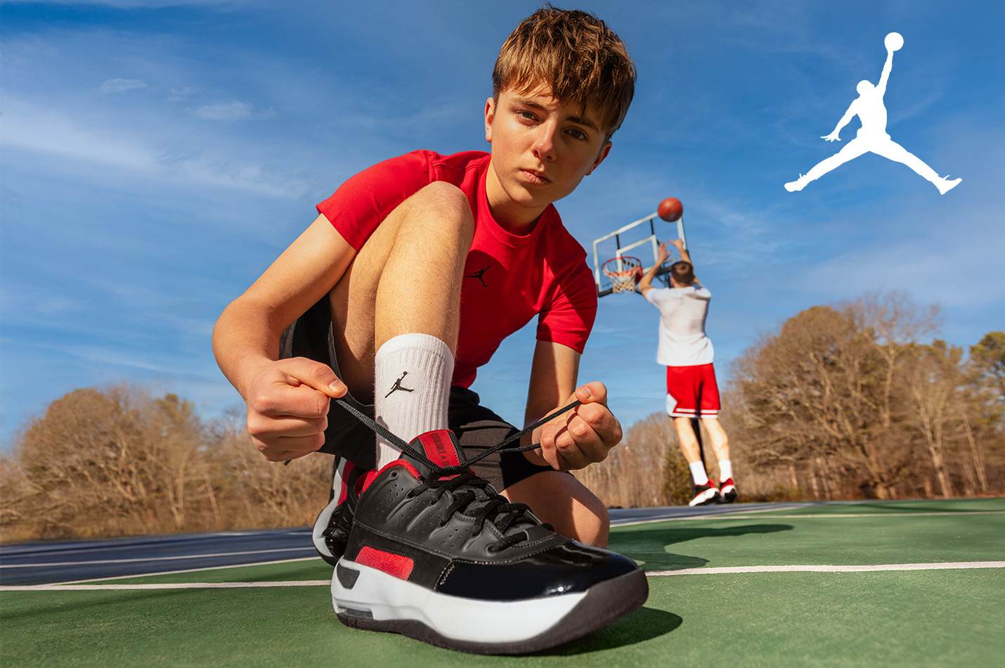 A teenage boy tying his Jordan Brand basketball shoes on a basketball court outside while a friend shoots a ball towards the hoop in the background.
